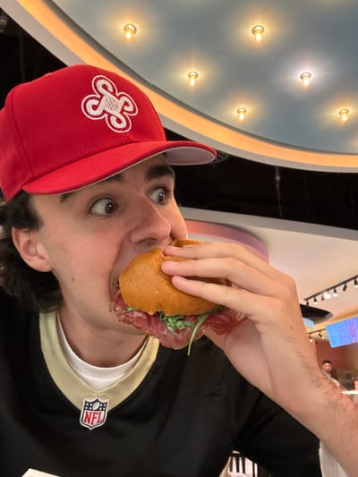 A person is eating a sandwich indoors, appearing to be in a restaurant or food establishment. The person is wearing a red baseball cap with a white clover-like logo and a black and gold shirt with an NFL logo. The background includes a wall with embedded lights and other patrons in the distance. Based on the hat and the shirt, it seems that the person is a New Orleans Saints fan. The city of Ferno, Italy cannot be determined from this image.
