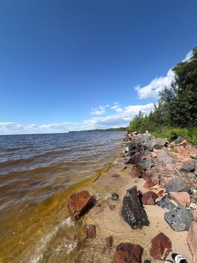 A wide-angle, eye-level, vertical shot depicts a rocky shoreline with a calm, brown-tinted body of water extending to the horizon under a clear blue sky dotted with white clouds. The foreground features a sandy beach with numerous large, dark gray and reddish-brown rocks, some partially submerged in the water. The water's edge laps gently against the sand and rocks, creating white foam. In the middle ground, along the rocky bank, a few people are visible, some sitting on the rocks and others standing, with a dense line of green trees forming the background. The overall scene suggests a sunny day at a natural waterfront location.