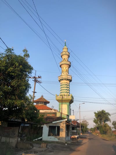 A tall, slender mosque minaret with green and white zigzag patterns dominates the center of the frame against a clear blue sky. The minaret is segmented, with several levels adorned with decorative elements and topped with a spire featuring a crescent moon emblem. Numerous power lines crisscross the sky, some draped low over the scene. To the left, a large, leafy green tree partially obscures a power pole with insulators and transformers. In the foreground and middle ground, several low-lying buildings with tiled roofs are visible, some appearing dilapidated. A paved road stretches into the distance on the right side of the image, lined with trees and more buildings, suggesting a semi-urban or rural setting. The lighting indicates it is late afternoon or early morning, with warm sunlight casting shadows. The scene appears to be in a residential area or along a road within a settlement in Besuki, Indonesia. There are no people clearly visible in the image.