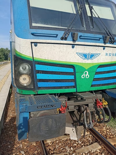 The image shows the front of a Siemens electric locomotive in Bliznets, Bulgaria, as evidenced by the Bulgarian Railways logo. The locomotive is painted in a blue, white, and green livery. The headlight on the left is illuminated. The buffer beam is visible along with coupling mechanisms. The train is positioned on a railway track alongside a concrete platform. Small stones cover the ground between the track and the platform.