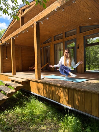 A young woman with long blonde hair, wearing sunglasses, a white long-sleeved top, dark blue pants, and white socks, is performing a dynamic balancing pose on a light blue yoga mat. She stands on her left leg with her right leg bent at the knee, foot near her left calf, and her arms slightly extended for balance. She smiles faintly, suggesting enjoyment.

The scene is set on a spacious wooden deck attached to a modern, multi-level wooden cabin or house, likely in Vozhoiskii, Russia. The building features large, angled windows that reflect the surrounding green trees and bright blue sky. The exterior walls are constructed from light brown wooden planks. Below the deck, lush green grass and various plants grow abundantly.

The sun is bright, casting distinct shadows across the wooden deck and on the vegetation below, indicating either morning or late afternoon. Decorative string lights with clear bulbs are strung along the wooden beams supporting the deck's roof, adding to the inviting ambiance. Through one of the cabin's large windows, a wooden chair is visible inside. The number "17" is affixed to the window frame near the woman. The overall impression is one of leisure and well-being in a natural, sunny environment.
