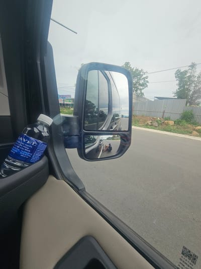 In Lương Sơn, Vietnam, a water bottle with a blue label reading "TRAVEL" and Vietnamese text is placed in a car's interior storage compartment. The bottle is positioned on the left side of the frame, leaning against the door panel. A side-view mirror reflects the road ahead, showing two individuals kneeling on the asphalt. The mirror is mounted on the vehicle's exterior, to the right of the water bottle. The road extends into the distance, bordered by a gray metal fence and vegetation on the right. The sky is overcast.