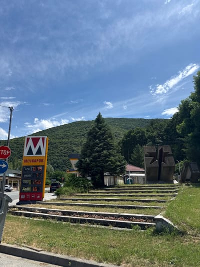 This image depicts an outdoor scene in Vetren Dol, Bulgaria, captured on a bright, sunny day with a clear blue sky dotted with wispy white clouds. The central focus is a tall, vertical gas station sign for "МЕЧКАРОВ" (Mechkarev). The sign displays current fuel prices in Cyrillic, likely in Bulgarian levs per liter: LPG at 0.75, Diesel at 1.71, A95H at 1.56, and A100 at 1.67. It also indicates availability of WC, WiFi, and acceptance of VISA payments.
In the foreground, partially obscured, are a red octagonal STOP sign and a blue circular directional sign with a white arrow. To the right, the ground slopes upwards with neatly arranged concrete terraces, filled with grass and some sparse vegetation. In the middle ground, substantial concrete structures, possibly part of a monument or architectural installation, are visible among dense trees. A building with a reddish-brown tiled roof can be seen behind these structures.
The background is dominated by a vast, rolling mountain range covered entirely in lush, dense green forest, extending to the horizon under the clear sky. The overall scene suggests a blend of natural landscape and roadside amenities in a mountainous region, under pleasant weather conditions.