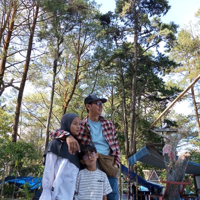 In the city of Gundaling 2, Indonesia, a family of three poses for a photograph amidst a lush forest of tall, mature pine trees. The family consists of a man, a woman wearing a hijab, and a young boy. The man, in his 40s, sports a plaid shirt, a blue t-shirt, eyeglasses, and a baseball cap. He has his arm around the woman's shoulders. The woman, who appears to be in her late 20s or early 30s, is dressed in a white shirt and a dark hijab, with a watch on her left wrist. The young boy, around 8 years old, is positioned in front of the couple, wearing a striped t-shirt and glasses. He looks towards the camera with a neutral expression. The background is dominated by the dense canopy of pine trees, with sunlight filtering through the branches, creating dappled light and shadows. In the middle ground, there are glimpses of some rudimentary structures, possibly part of a recreational area or a local attraction, including blue tarpaulins and wooden supports attached to a tree trunk adorned with colorful paint. The overall atmosphere is casual and relaxed, suggesting a family outing in a natural setting.
