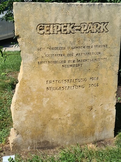 A weathered, light-colored stone monument stands upright in a grassy area with some green foliage and a portion of a white car visible in the background. The monument has an irregular shape with a chipped left edge. Engraved text appears on the front of the stone. The top line is in large, bold, sans-serif capital letters reading "GEIPEK-PARK". Below this, smaller engraved text lists acknowledgements and dates. The text reads: "DEM FÖRDERER U. GÖNNER DER VEREINE GESTALTER DER PARKANLAGEN EHRENBURGER DER MARKTGEMEINDE NEUMARKT ERSTGESTALTUNG 1904 NEUGESTALTUNG 2004". The monument is situated outdoors, likely in a park or public space. The lighting suggests it is daytime, with the sun casting shadows and highlighting the textures of the stone.