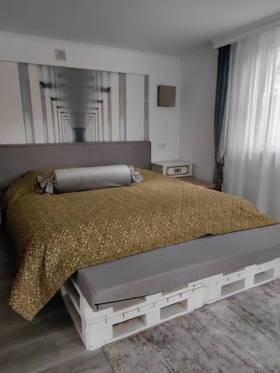 A bedroom is shown with a bed that has a grey upholstered headboard and a gold patterned comforter with a bolster pillow. The bed frame is made of white painted wooden pallets. A grey upholstered bench sits at the foot of the bed, also made from pallets. A large mural of a corridor with receding pillars is on the wall behind the bed. To the right of the bed, sheer white curtains hang over a window, with a dark blue curtain panel to the left of them. A small white nightstand with gold trim is next to the bed. The flooring appears to be light-colored wood planks with a patterned rug in front of the bench. A square metallic object is mounted on the wall to the right of the mural. The scene is indoors in a room that appears to be part of the Алесь-2002 residential complex in Russia.