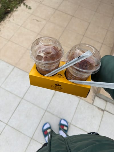 A top-down shot shows a person holding a yellow cardboard carrier containing two iced coffee drinks. The drinks are in clear plastic cups with dome lids and straws. The carrier has the words "ΓΑΤΙΑΔΗΣ FRESH" printed on the side. The person is wearing a green jacket and slides with blue, white, and red detailing. The setting appears to be an outdoor paved area, possibly in Megas Alexandros, Greece, based on the brand name on the carrier.