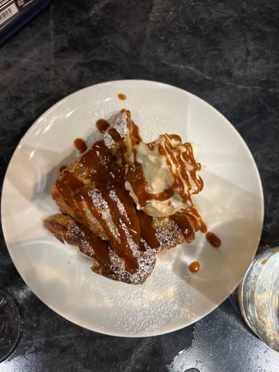 A white, circular ceramic plate holds a dessert or breakfast item, likely French toast or a similar pastry, centrally on a dark, mottled countertop. The dish features several golden-brown pieces, heavily coated with a rich, amber-colored caramel sauce and generously dusted with white powdered sugar. A distinct dollop of white whipped cream or ice cream rests on the upper right portion of the main item, also drizzled with the same caramel sauce. In the upper left background, a dark blue object with a white label showing partial text and a barcode is visible. A portion of a reflective, metallic or glass drinking vessel is present in the lower right foreground. The scene is indoors, likely within a restaurant or cafe located in Orléans, France.