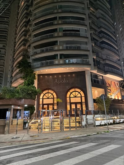 An upward-facing image depicts a tall building with a curved facade. Its entrance is labeled "Apolo" above arched doorways, illuminated from within. An adjacent street showcases reflective metal stairs and a crosswalk. The building has curved balconies and dark windows. Lush greenery borders the base of the building near the street. A painting adorns the wall above the main entrance.