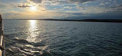 The image captures a wide, calm body of water with gentle ripples, reflecting the golden sunlight of a sunset. The sun, partially obscured by dramatic clouds, casts a bright, shimmering path across the water's surface. In the distance, a dark, tree-lined horizon is visible, with faint mountain silhouettes forming the backdrop under a partly cloudy sky. On the left side of the frame, a railing of a vessel, likely a boat or ferry, is partially visible, suggesting the perspective is from a moving craft. The scene evokes a serene and tranquil atmosphere of a coastal or lakeside landscape near Pristan'-Przheval'sk, Kyrgyzstan.
