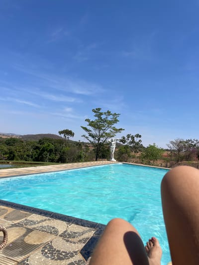 The image presents a first-person, low-angle view from a seated position, looking out over a swimming pool towards a rural landscape under a clear blue sky, located near Pirenópolis, Brazil.
In the immediate foreground, the lower legs and feet of an individual are visible, resting. The skin is light-toned, and the toenails on the visible foot are painted a reddish-pink color. Below the legs, the dark-tiled edge of a swimming pool is visible, featuring a geometric pattern in black, grey, and white.
The central subject is a rectangular swimming pool filled with clear, turquoise-blue water, reflecting sunlight and showing subtle ripples. Beyond the pool's edge, a light-colored stone paving is present. At the far end of the pool, slightly to the right of the vertical center, stands a tall, white, classical-style female statue, draped, with her arms slightly raised.
Behind the statue and pool, a vibrant green landscape unfolds. A prominent, leafy tree with a relatively slender trunk is positioned directly behind the statue. Further back, the terrain consists of rolling hills densely covered in various shades of green foliage and trees, characteristic of the Brazilian cerrado or Atlantic Forest transition zone. A small, dark-colored body of water, possibly a pond or lake, is visible on the far left side of the background, nestled among the trees.
The sky occupies the upper half of the frame, exhibiting a deep, unblemished blue with only a few faint, wispy white clouds, indicating bright, sunny weather. The overall scene depicts a serene outdoor leisure environment within a natural, hilly setting.