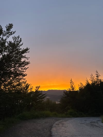 The image depicts a tranquil sunset scene on a rural dirt road in Strazha, Bulgaria. Silhouetted trees line the foreground and frame a vibrant orange and yellow sky. In the distance, rolling hills and mountains are visible, softened by a light haze. The road itself is made of gravel and asphalt, with some cracks visible on the paved section. The overall atmosphere is serene and peaceful.