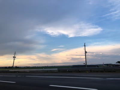 The image shows a landscape scene on a cloudy day, likely during twilight. A paved road with white lane markings dominates the foreground. A metal guardrail separates the road from a field in the middle ground. Beyond the field, there are power lines supported by utility poles. In the distance, there are some buildings and trees, suggesting a rural or semi-rural setting. A Canadian flag is visible on a pole behind the guardrail. The sky is filled with various cloud formations, with patches of blue sky and warmer tones of yellow and pink indicating the setting sun. The overall mood of the image is calm and serene, with a hint of the approaching evening. There is no visible text in the image.