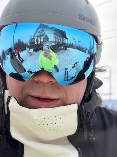 The image presents a close-up, first-person view of an individual wearing winter ski gear. The primary subject is wearing a grey ski helmet with "SINNER" emblazoned on the side, and reflective blue mirrored ski goggles. The lower part of the subject's face, including the nose, mouth, and stubbled chin, is visible, partially covered by a white and black neck gaiter featuring a perforated pattern. The subject's black and white ski jacket is also visible.

The blue mirrored goggles reflect a panoramic winter scene, identified as Novoabzakovo, Russia. The reflection clearly shows a two-story white building with dark wooden trim and a dark roof, resembling a ski lodge, situated on a snow-covered slope. In the immediate foreground of the reflection, a person dressed in a bright neon yellow ski suit and a light-colored helmet is visible, holding a silver smartphone to take a selfie. Other individuals are present on the snowy terrain: one person in a red jacket on a snowboard, and another in a beige jacket. Ski equipment, including skis and poles, are discernible on the snow. Ski lift infrastructure is visible in the background against a light blue sky. The environment is a bustling ski resort setting.