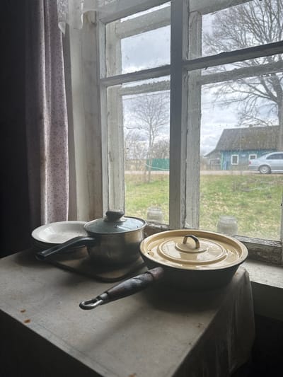 The image depicts a cluttered scene inside a building, potentially a house. A weathered window frame with multiple panes is visible, revealing an exterior view of an overcast day with bare trees, a green field, and a house, possibly with a vehicle parked in front. On a surface next to the window, there is a stack of cookware, including a tan-colored lid, a saucepan with a lid, and a white plate. The surrounding area is dimly lit, and the surfaces appear aged and worn. A curtain with a floral pattern hangs below the window. This scene is likely located within Студзёнкаўскі сельскі Савет, Belarus, based on provided location information.