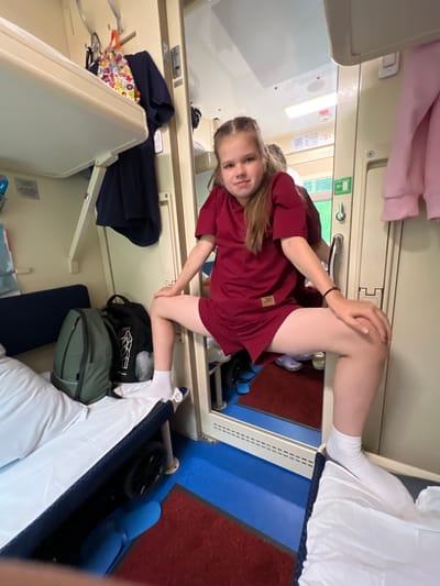 A young girl with blonde hair braided into two pigtails is positioned in a narrow train compartment, leaning forward and resting her elbow on her knee. She is wearing a short-sleeved, dark red t-shirt and white socks. Her expression is a neutral gaze towards the camera.
The compartment appears to be a sleeping berth on a train, with bunks visible above and to the side. The walls are a neutral beige color, with metal trim and door handles. A pink garment is draped over the upper bunk. On the lower bunk, a blue bag and a green backpack are placed. White linens cover the sleeping areas. There is a blue plastic seat in the corner. The floor is carpeted with a dark red material and has a blue accent strip.
The overall scene suggests a travel or a stopover in a train compartment. The lighting appears to be artificial, typical of an indoor train environment, making it difficult to determine the time of day or weather outside. There is no visible text in the image.