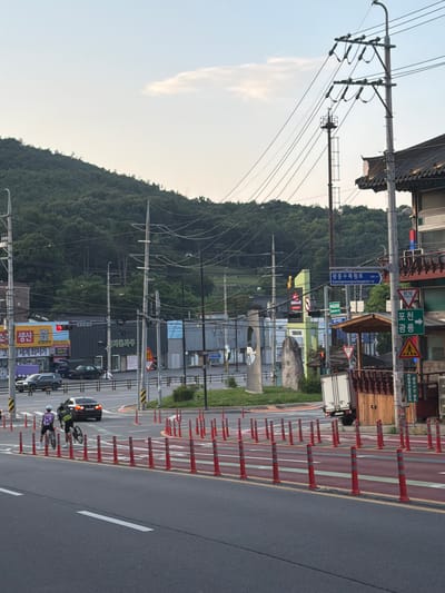 The image depicts a street scene in Pocheon-si, South Korea, under a partly cloudy sky. A two-lane road occupies the foreground, with a bike lane marked by red and white bollards. Two individuals on bicycles are visible in the bike lane. Across the street, several buildings are present, including a traditional-looking structure with a tiled roof on the right. Numerous utility poles and overhead wires crisscross the scene. Signage, including traffic signs in Korean, is visible on poles and buildings. A large hill covered in trees forms the backdrop. Traffic is light, with a few cars and a delivery truck visible.