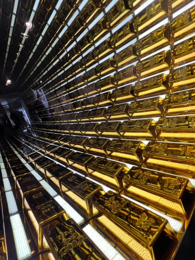 The image shows a large display of gold bars arranged in rows, creating a tunnel-like effect. The gold bars are highly reflective and appear to be lit from behind by bright white lights. Some of the bars have markings indicating "FINE GOLD" and weight. In the background, there are glimpses of people and what appears to be a dark, reflective environment. The perspective suggests a wide-angle shot, looking down a corridor or through an expansive display.

This display of gold bars is located in the S.E.M.S. (Société d'Economie Mixte Maritime et Portuaire) building in Honfleur, France. The building, which houses the port authority, features a unique architectural element where the main staircase is lined with these gold bars as a decorative and symbolic element representing the wealth and importance of the port.