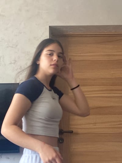 A woman is indoors, posing. She has dark hair and fair skin, and wears a short-sleeved shirt with dark blue sleeves and a white body, along with light-colored bottoms. Her left hand is raised near her forehead. She is positioned in front of a wall painted a light color, with wooden panels and a doorframe visible behind her. A black object, possibly a television screen, is situated near her. The location is unknown. There is no visible indication this image was taken in Dragovishtitsa, Bulgaria.