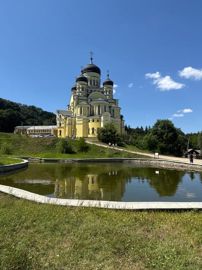 This is a wide-angle, eye-level, outdoor photograph taken on a sunny day in Ciuciuleni, Moldova. The image features a large, yellow orthodox church with multiple black domes and crosses, situated on a grassy hill. In the foreground, a curved pond with green, reflective water is bordered by a white stone edge and surrounded by green grass. The church is the dominant subject, with its intricate architectural details visible. Several people are walking on a path leading to the church, and a few cars are parked on a road further back on the hillside. The background consists of a dense forest and a clear blue sky with scattered white clouds. The overall scene evokes a sense of peace and architectural grandeur within a natural setting.