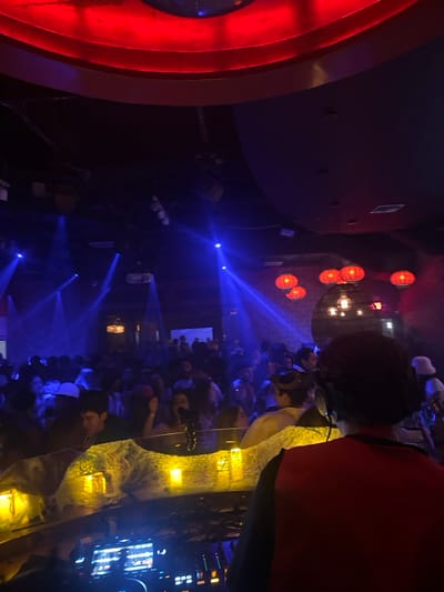 A dimly lit indoor venue, potentially a nightclub or concert hall, is filled with a large crowd. Red spherical lights and modern chandeliers illuminate the scene. Blue laser lights cut through the air, adding to the vibrant atmosphere. Sections of red decor are present on the left. The crowd seems dense and mostly facing the same direction, possibly toward a stage or performance area. The image appears to be taken from a high vantage point.