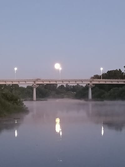 The image depicts a calm river at dusk, with a thick layer of fog hovering just above the water's surface. A concrete bridge spans across the river in the midground. The bridge is illuminated by several streetlights, two of which are particularly bright and appear to be reflecting in the water below. The riverbanks are lined with dense green foliage, suggesting a natural or semi-natural setting within a city, possibly in the vicinity of Buy, Russia, given the provided context. The sky is a pale blue-gray, typical of twilight. There are no people or vehicles visible in the scene. The overall atmosphere is serene and quiet.