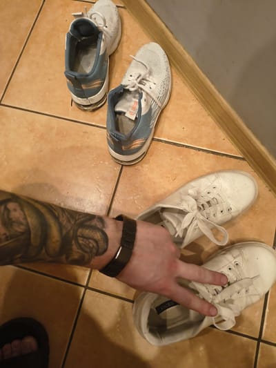 The image shows a close-up view of several pairs of athletic shoes and a person's arm with a tattoo on a tiled floor. Two pairs of white and blue athletic shoes are positioned in the upper left and upper right quadrants of the image, with one shoe from each pair appearing to be worn, revealing a blue and orange interior. In the lower half of the image, a person's arm with a dark band on the wrist is visible, displaying a tattoo of a woman and a snake. The person's hand is extended over a pair of white leather sneakers, with two fingers forming a peace sign. Another white sneaker is visible in the bottom right corner. The floor is tiled in a warm, earthy tone with dark grout lines. The background consists of a wall with a light brown baseboard. There is no information in the image to indicate a location in Grójec, Poland.