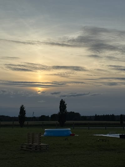The image depicts a rural outdoor scene in Jeedasküla, Estonia, during what appears to be twilight. The sky is filled with a variety of clouds, with the sun partially obscured by them, casting a soft glow. In the foreground, a grassy field is visible, with several objects scattered across it. To the left, there is a structure made of wooden pallets. In the middle ground, a blue inflatable pool is present, and to its right, a white tarp or pool cover is laid out. Beyond the immediate field, a line of trees and vegetation forms the horizon, with a power line visible in the distance. The overall atmosphere is serene and quiet, suggesting a peaceful evening.