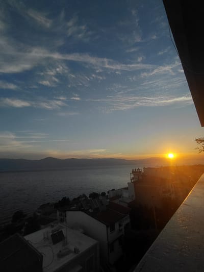 The image depicts a sunset over a body of water, with a coastal town visible on the hillside. The sky is a gradient of blues and oranges, with streaks of white clouds. The sun is a bright orb, casting a warm glow over the scene. In the foreground, a balcony railing is visible, partially obscuring the view. The town consists of white buildings with red-tiled roofs, nestled on the slope. Mountains form a silhouette in the distance across the water. The image appears to have been taken from a high vantage point. The location is identified as the Municipal Unit of Aegio, Greece.