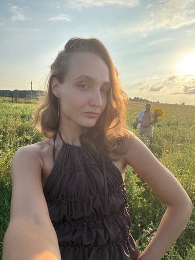 A young woman in a brown ruffled dress is taking a selfie in a field of tall grass and wildflowers, with a man in the background holding a large bouquet of sunflowers. The sun is low in the sky, casting a warm glow over the scene. The woman's hair is light brown and wavy, and she is wearing a long, delicate earring. Her expression is neutral as she looks at the camera. The man is wearing a light blue unbuttoned shirt and tan shorts. He appears to be walking towards the woman. The landscape is rural, with power lines and trees visible in the distance. The location is identified as Aksaitovo, Russia.