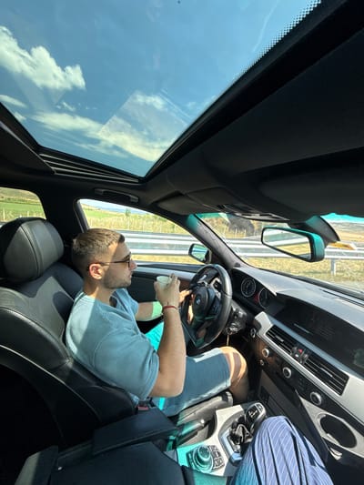 In the city of Kermen, Bulgaria, a man is captured from the chest up while driving a car. He is wearing sunglasses and a light blue striped t-shirt, with turquoise shorts. In his right hand, he holds a white paper cup, and his left hand is on the steering wheel. The car's interior is visible, featuring a black leather seat, a sunroof with a view of a blue sky with white clouds, and a dashboard with gauges and a car stereo. The surrounding environment appears to be a highway with green fields and trees visible through the car window. The image is taken on a sunny day.
