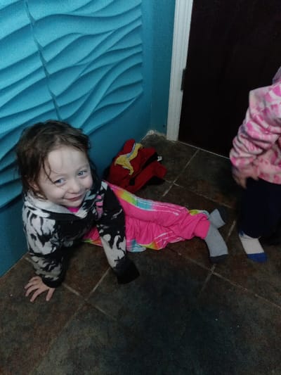 A young girl with dark hair, wearing a black and white hooded shirt and tie-dye pants, poses on a tiled floor against a blue textured wall. She is smiling at the camera. Next to her are some red and yellow clothes. Another child, wearing a pink patterned shirt and dark pants, stands partially in the frame on the right. A dark door is visible in the background.