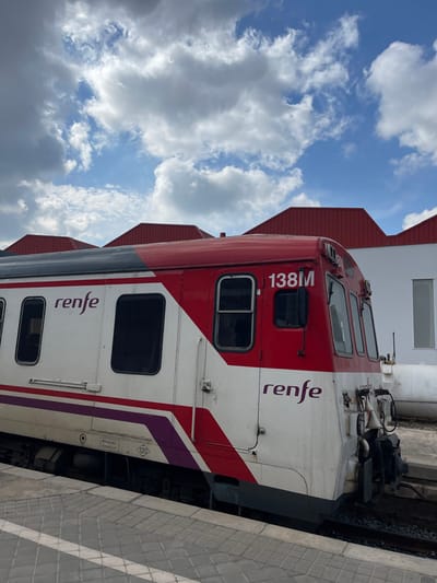 A stationary RENFE train is depicted at a railway station or siding in Buñol, Spain. The train unit, predominantly red and white with a purple stripe along its lower side, displays the "renfe" logo prominently on its side and the designation "138M" on the front cab section. The lower body of the train exhibits wear and dirt. It rests on a set of tracks adjacent to a grey-tiled platform. In the background, a white industrial-style building is visible, with multiple red, gabled roofs extending above it to the left. A large white horizontal pipe lies on the ground behind the train, near the white building. The sky is partially cloudy with significant white cumulus clouds and patches of clear blue. No individuals are present in the scene.
