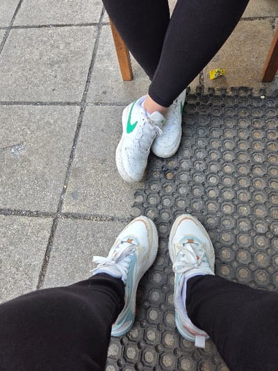 An overhead view captures the lower legs and feet of two individuals seated in an outdoor area in Gevgelija, North Macedonia.

The upper subject wears black leggings and white athletic shoes, each featuring a prominent green Nike 'swoosh' logo on the lateral side. Their legs are crossed at the ankles. A segment of a light brown wooden chair leg is visible to the right of their left foot.

The lower subject wears black leggings and white athletic shoes accented with light blue and beige elements. Their feet are positioned parallel on the ground, slightly separated.

The ground surface is divided into two distinct sections: grey concrete pavement with visible seam lines on the left, and a black rubber mat with a repeating circular/octagonal pattern covering the right side. A small, crumpled yellow object, consistent with a wrapper, is present on the black mat between the two subjects' footwear. The exact location within Gevgelija cannot be determined from the visual information provided.