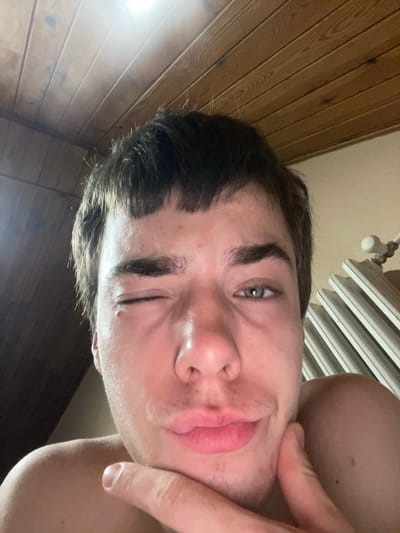 A close-up selfie shows a young person with short, dark hair and a light complexion. Their left eye is partially closed, while the right eye is open, revealing a green iris. Their lips are parted, and their right hand is resting on their chin. The background consists of a wooden ceiling and a white radiator with a valve. The lighting suggests an indoor setting. The image does not contain any specific information about the city of Jihlava, Czechia.