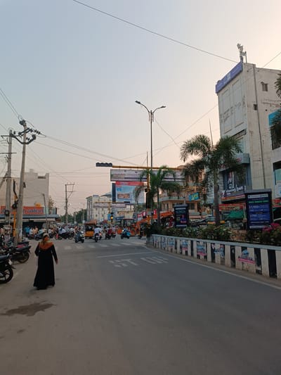 The image presents an urban street scene in Anantapur, India, observed under a clear, pale sky with a slight warm tint. The foreground features an asphalt road with a painted "STOP" marking. One individual, dressed in a long black garment and an orange-brown head covering, walks away from the camera on the left side of the road.

To the left, a row of motorcycles and scooters are parked along the curb, fronting multi-story commercial buildings partially obscured by numerous utility poles and an intricate network of overhead power lines. Visible signage includes "శ్రీ లక్ష్మి ఫ్యాన్సీ హోటల్" in Telugu.

The main roadway displays moderate to heavy traffic, predominantly consisting of motorcycles, scooters, and auto-rickshaws. A horizontal traffic signal, emitting a yellow light, is positioned over the road in the mid-ground. A central median, constructed from a black and white striped concrete barrier, is visible, adorned with various posters and supporting palm trees and flowering plants.

On the right side of the street, beyond the median, multiple multi-story commercial buildings are present. A prominent, light-colored building on the far right features cellular antennas on its roof and a blue sign displaying "MAXLIFE" alongside Telugu script. Further commercial signs are visible, including "Anantapur's NextGen," "Unacademy," and a "Tiffin & Biryani Center." A yellow construction vehicle is visible on the right side of the road near the intersection.