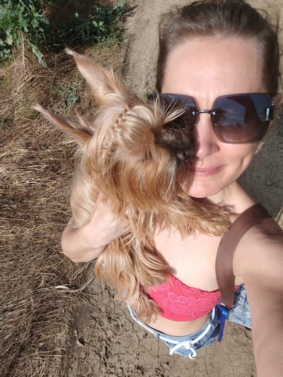 A woman is holding a Yorkshire Terrier while standing outdoors. She is wearing sunglasses, a pink lace bralette, and denim shorts. The ground appears to be dry and cracked, with some dry vegetation and green plants in the background. There is no indication of the location in Fyodorovskaya, Russia in this image.