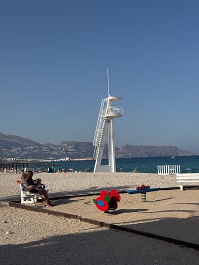 This is a photograph taken on a sunny day at a beach in l'Alfàs del Pi, Spain. The foreground features a sandy area with a children's playground that includes a red and blue spring rider. A woman and a child are seated on a white bench to the left of the playground. Beyond the playground, a white lifeguard tower stands prominently on a pebbled beach. The background shows the turquoise Mediterranean Sea, with mountains and coastal buildings visible in the distance. Several beach umbrellas, lounge chairs, and people enjoying the beach are scattered further down the shoreline. The sky is clear and blue.
