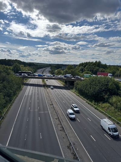 The image captures a view of a multi-lane highway in Dartford, United Kingdom, with traffic moving in both directions. The highway is bordered by lush green trees and vegetation. Above the highway, a bridge supports traffic, with vehicles visible on it as well. The sky is partly cloudy, with dramatic white and grey clouds against a blue backdrop. Several vehicles are present on the highway, including cars, vans, and a bus. Some traffic appears to be moving steadily, while other sections show congestion. The lighting suggests it is daytime, possibly late morning or afternoon. The perspective is from an elevated position, looking down onto the highway.