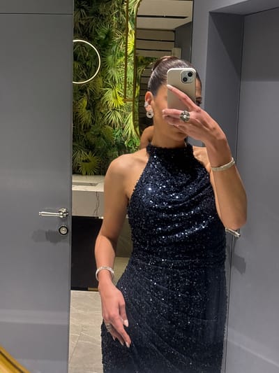 A person stands in a bathroom, taking a selfie in a mirror. They are wearing a sleeveless, sequined dress. The person is holding a white smartphone with the camera visible, obscuring their face. On their left wrist is a bracelet, and a ring on their finger. The bathroom has a green decorative wall with a circular light. A tap and sink fixture are visible. The location is presumed to be in Masis, Armenia.