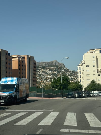 The image captures a street scene in Calp, Spain, with a clear blue sky suggesting a sunny day. The perspective is from a low angle, looking upwards and to the right. Dominating the scene are several multi-story buildings. To the left, a tall, light-colored apartment building with numerous windows and balconies is visible. Behind it, a densely packed hillside settlement with white houses and red-tiled roofs stretches towards a rocky mountain. In the foreground on the right, a street with a pedestrian crossing is visible. A checkered blue and white van is parked on the street. In the background, a line of parked cars and trees are seen along the roadside, with more buildings and infrastructure further up the hill. The architecture suggests a coastal Mediterranean town. No people are clearly visible, but the presence of vehicles and buildings indicates an urban or semi-urban environment. The time of day appears to be daytime, given the bright lighting and shadows. The overall impression is a snapshot of a residential area in a Spanish coastal town. The van has visible text: "BIMBO" and a license plate ending in "66 MNP".