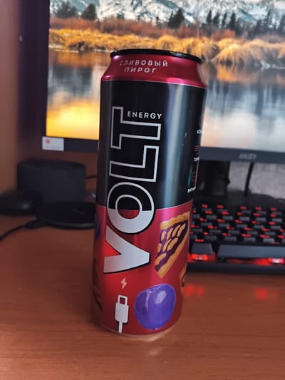 A can of VOLT energy drink, with a red and black design, is positioned in the foreground, angled slightly towards the viewer. The can features Russian text, including "СЛИВОВЫЙ ПИРОГ" (plum pie) and "ENERGY". A graphic of a slice of pie and blueberries is also visible on the can. To the right of the can, a portion of a black computer keyboard with illuminated red keys is present. In the background, a computer monitor displays a landscape image of a body of water with trees and mountains. The scene is set on a wooden surface. There is no indication of the location being Mokshan, Russia.