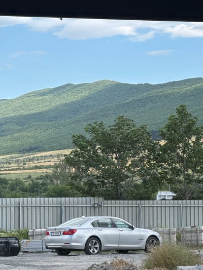 The image depicts a silver sedan parked next to a corrugated metal fence. The car is facing towards the right of the frame and its rear is visible. The license plate reads "MA 454 RK". Behind the fence, there is lush green foliage and rolling hills covered in dense vegetation, suggesting a natural landscape. The sky is a clear blue with a few scattered white clouds. The ground where the car is parked appears to be a gravelly or unpaved surface. The overall scene suggests an outdoor setting, possibly in a rural or semi-rural area. The lighting indicates it is daytime, likely with bright sunshine given the clear sky. The perspective is from a slightly elevated position, looking down at the car and the landscape. There is no visible activity or people in the frame. The image has a slightly tilted orientation.