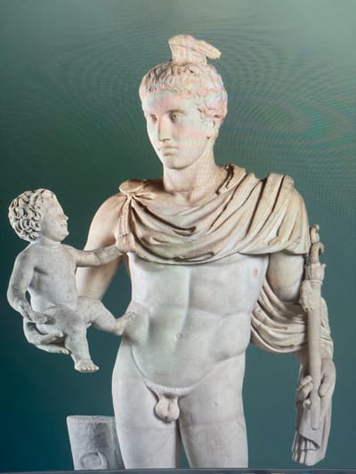This is a photograph of a marble sculpture depicting a male figure holding a baby. The sculpture is oriented vertically, with the figures appearing to be lying down. The male figure is muscular and draped in flowing fabric that covers part of his torso and upper legs. He has short, curly hair styled in a topknot. His head is tilted downwards, and his gaze appears to be directed at the baby.

The baby is held in the male figure's arms and is also depicted as muscular. The baby has curly hair and its legs are bent. The sculpture is rendered with a high degree of detail, showcasing the musculature of both figures and the texture of the drapery.

The background is a textured, muted green, suggesting a neutral display setting rather than a specific environment. There are no discernible indicators of time of day, weather, or specific location cues within the sculpture itself or its immediate surroundings. The overall mood conveyed by the sculpture is one of tenderness and care, as suggested by the embrace between the figures. There is no visible text in the image. The location context provided is Kafr Yasif, Israel, but this information is not directly evident from the image itself.