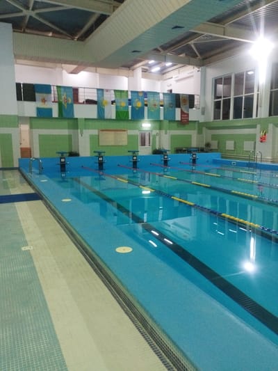 The image shows the interior of a swimming pool facility, likely a public or training pool. The main subject is a clear blue swimming pool with multiple lanes marked by ropes and buoys. Starting blocks for swimmers are visible at one end of the pool. Overhead, the ceiling features exposed beams and lighting fixtures, with some evidence of ventilation systems.

Along the wall above the pool, several flags are displayed, suggesting an event or a connection to different regions or countries. The walls are painted in a combination of green and white. A life vest in an orange and yellow color is hung on the wall to the right. Large windows are visible on the far wall. The floor area around the pool appears to be tiled with a patterned surface.

The location is identified as Қотырбұлақ, Kazakhstan. However, based solely on the visual information within the image, it is not possible to definitively confirm this specific location without external context or identifying markers within the scene itself. The visual elements are typical of an indoor swimming pool. There are no people visible in the image.
