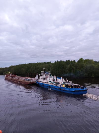 The image depicts a blue and white tugboat pulling a rust-colored barge on a river in Volokov Most, Russia. The sky is overcast and cloudy. The river is dark and has visible ripples and a wake trailing from the tugboat. Both banks of the river are lined with green trees, forming a dense forest in the background. The tugboat is attached to the barge at the front. There's some smoke coming from the vessel.