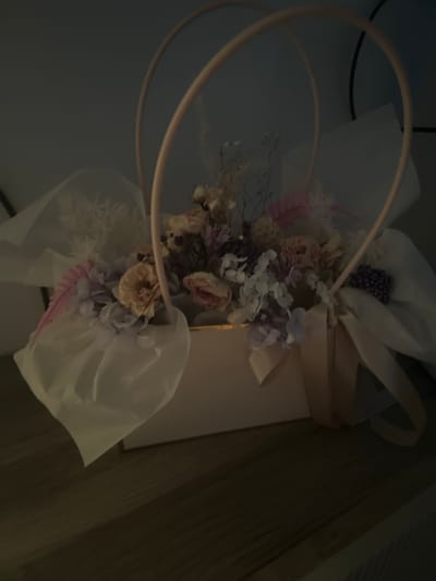 A bouquet of dried flowers is arranged in a light pink rectangular box with a gold trim. The bouquet contains various dried flowers in shades of white, cream, pink, and purple, along with some feathery accents and small, dark berry-like elements. A light tan ribbon is tied around the front of the box. Two curved handles, also light pink, extend from the sides of the box, arching over the bouquet. The box is placed on a wooden surface. The background is dark and appears to be a wall. The lighting is dim, casting shadows and making some details difficult to discern. There are no people or animals visible in the image. The image does not contain any information or context related to the city of Baymak, Russia.