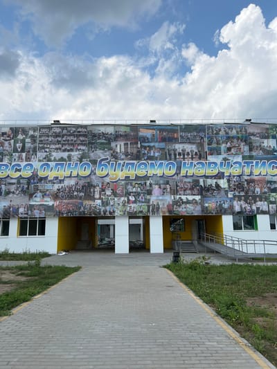 A modern, multi-story building facade, located in Staryi Saltiv, Ukraine, is depicted under a partly cloudy sky. The upper two-thirds of the building's front is covered by a large, panoramic banner or mural. This banner is a collage of numerous individual photographs, primarily depicting groups of people, likely students, engaged in various educational, social, and cultural activities. Prominently displayed across the center of the banner is text in Cyrillic, "ВСЕ ОДНО БУДЕМО НАВЧАТИСЬ," rendered in large blue letters with a yellow outline, set against a background that incorporates yellow and blue hues.
The lower portion of the building features a facade with white and bright yellow panels. A central recessed entrance, marked by large white pillars, appears dark and somewhat obstructed within, revealing internal debris or construction materials. White-framed windows are visible on the lower left and right sections of the building. On the right side, a set of concrete steps and an adjacent metal-railed accessibility ramp lead to an elevated entry point.
In the foreground, a gray paved walkway, composed of rectangular pavers and bordered by yellow lines, extends directly towards the building's central entrance. Areas of unkempt green grass and weeds flank both sides of the walkway. No living individuals are visible in the scene.