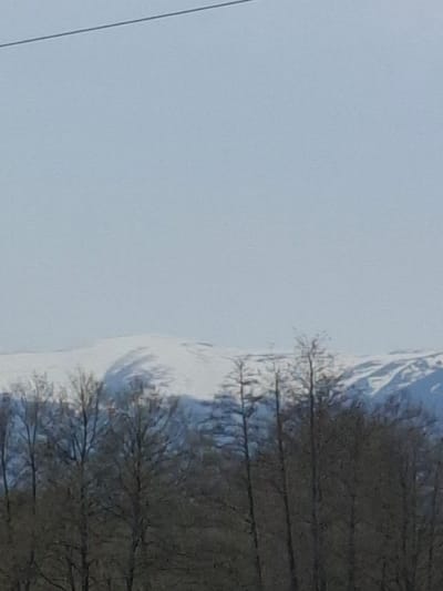 The image shows a landscape with a pale, overcast sky occupying the left portion. To the right, a snow-covered mountain range is visible, with bare trees standing in front of the snow. A power line is visible in the lower left corner. The location is presumed to be in Komarevo, Bulgaria, based on the prompt. No people, specific objects, actions, or interactions are discernible.
