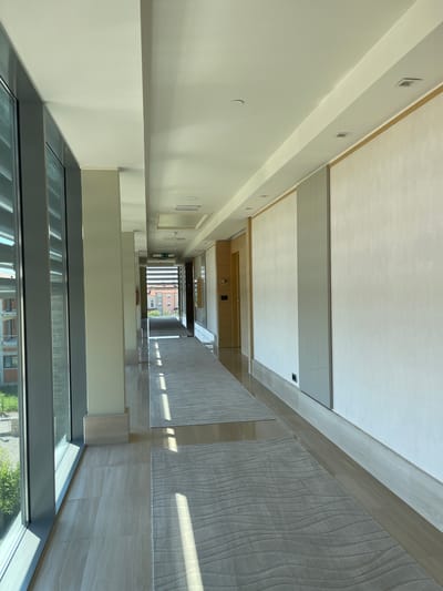 This media file captures an interior view of a modern hallway, likely in a building under construction or recently renovated, as indicated by the clean, unfinished elements and general sparseness. The perspective is from an elevated position looking down a long, wide corridor.

The hallway features a light-colored ceiling with recessed lighting and air vents, a pale yellow wall on the left, and a contrasting textured carpet and tiled floor on the right. The overall aesthetic is minimalist and contemporary.

Visible objects include doors on the left wall, some with small windows or panels, and what appear to be light fixtures and possibly electrical outlets. Towards the end of the hallway, a large window or glass partition offers a glimpse of an exterior scene with buildings and greenery, suggesting an urban or suburban setting. The time of day appears to be daytime, with sunlight casting shadows and highlights on the textured carpet.

No people are visible in the media file. The scene is static and devoid of any discernible activity. There is no visible text that can be clearly deciphered. The environment appears to be an indoor public space, possibly a hotel, office building, or residential complex, given the design elements. The location context provided is Crveni Vrh, Croatia, which adds a geographical dimension to the setting.